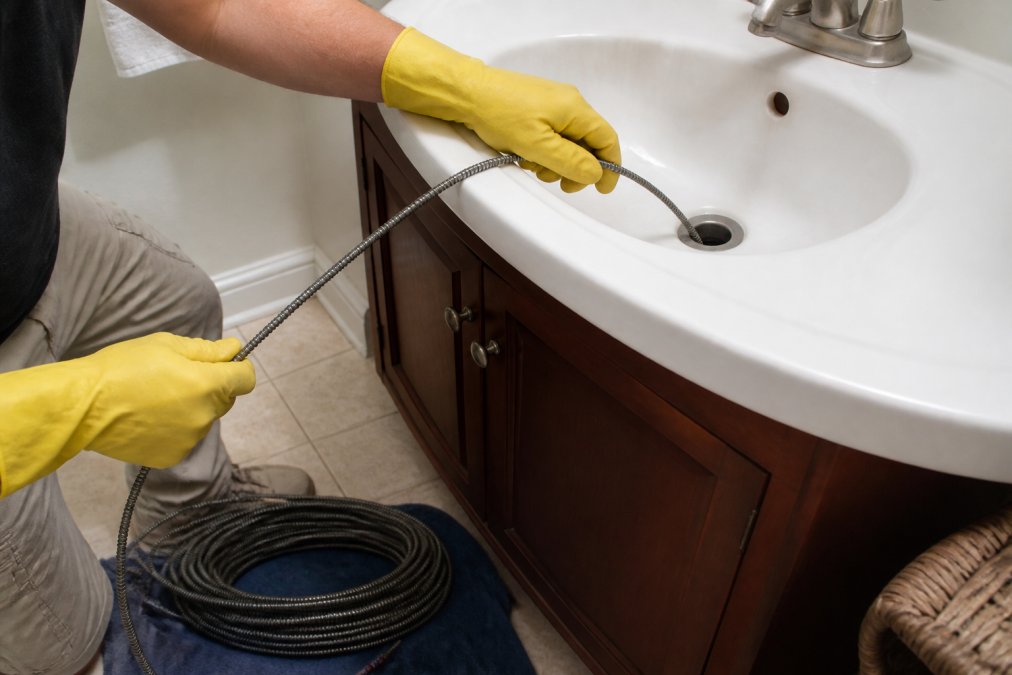 A person wearing yellow rubber gloves feeding a coiled metal drain snake into a bathroom sink drain, with the remaining snake coiled on a towel on the floor beside the vanity cabinet under bright bathroom lighting