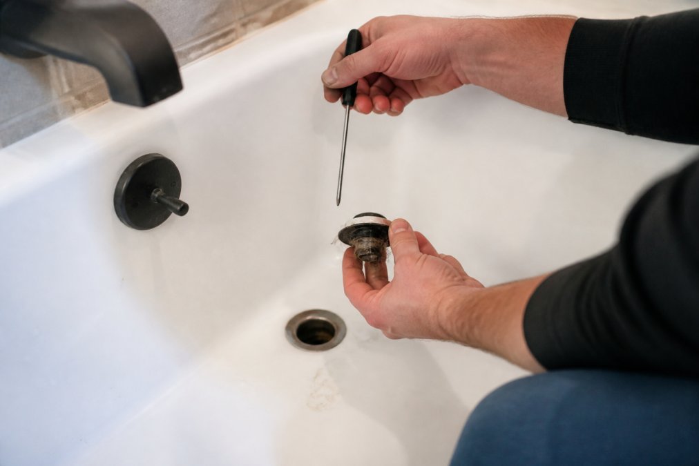 Homeowner using a screwdriver to remove a hair-clogged stopper from a slow draining bathtub drain.
