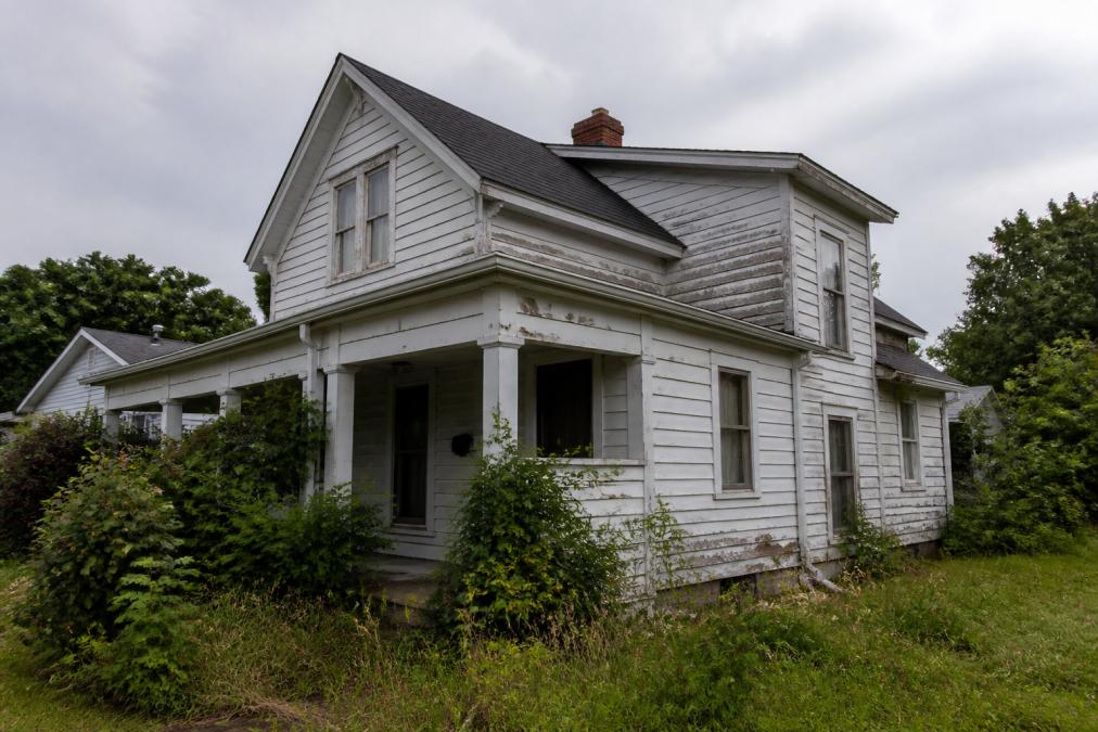 A home exterior showing visible wear with weathered siding, peeling paint, and overgrown landscaping representing an as-is property condition, overcast sky