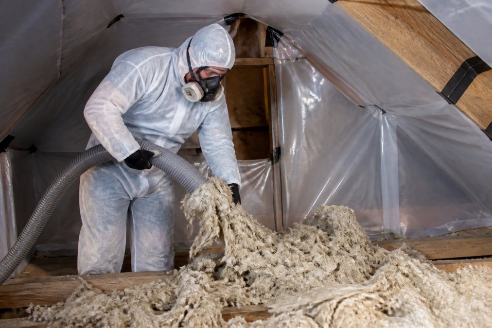 Professional wearing full protective equipment including respirator, gloves, and protective suit carefully removing insulation from loft space, with plastic sheeting containment visible around the work area