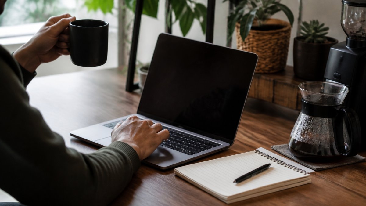 Hands holding a coffee mug while using a laptop to manage subscription settings in a cozy home office with coffee brewing nearby