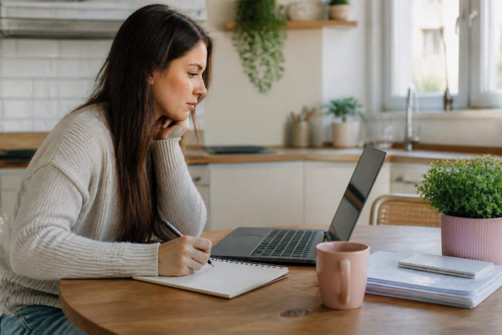 Woman at a kitchen table using a laptop and notebook to compare insurance quotes via a side-by-side process chart