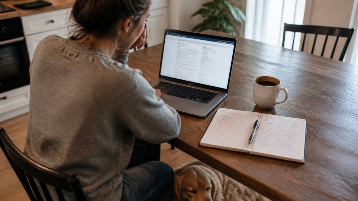 A pet parent sitting at a kitchen table with a laptop open to a shipping policy page, a notebook with handwritten cost comparisons, a coffee mug, and a dog resting at their feet