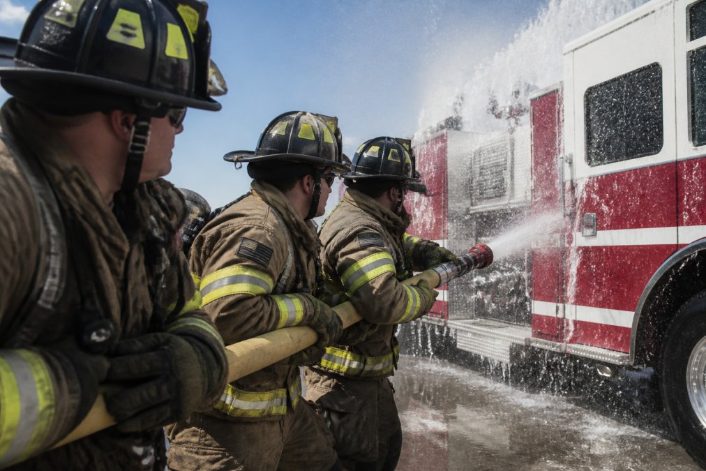 Close-up of firefighters holding a charged hose line with proper grip and positioning, water streaming over a fire engine during a wetdown ceremony, focused intensity and control evident in their stance