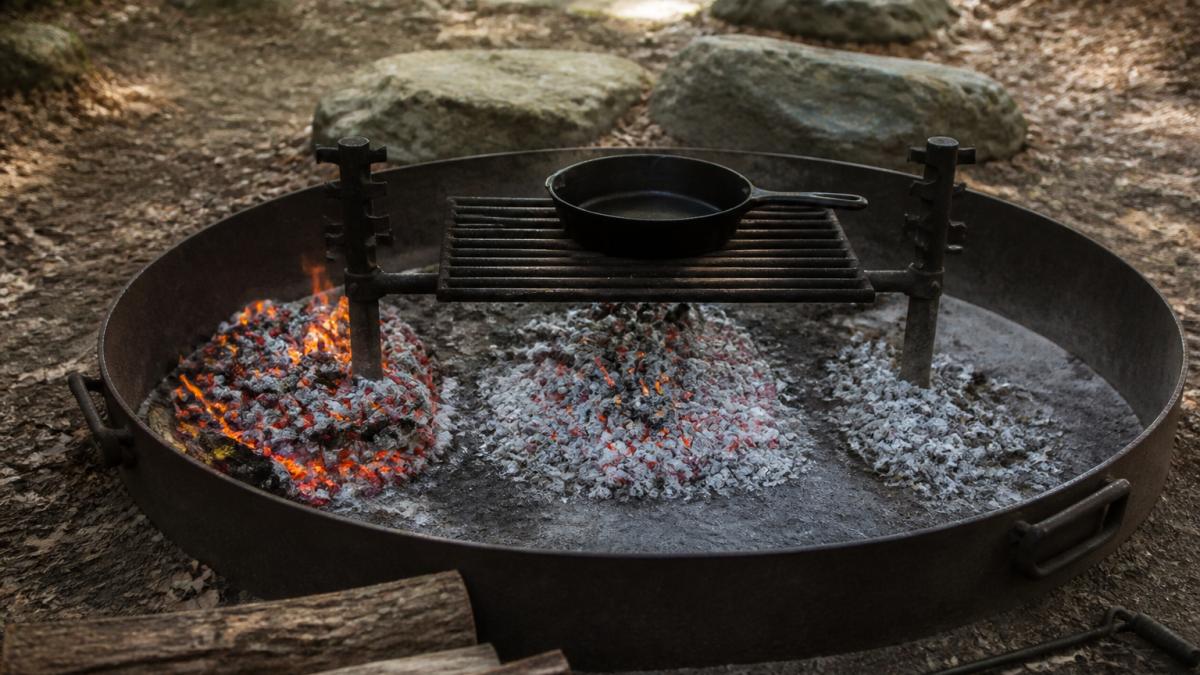 A campfire with hot coals raked into three distinct zones, a high-heat pile on one side, a medium-heat spread in the center, and a low-heat area with sparse coals on the other side, with a cast iron skillet sitting on a grate over the medium zone