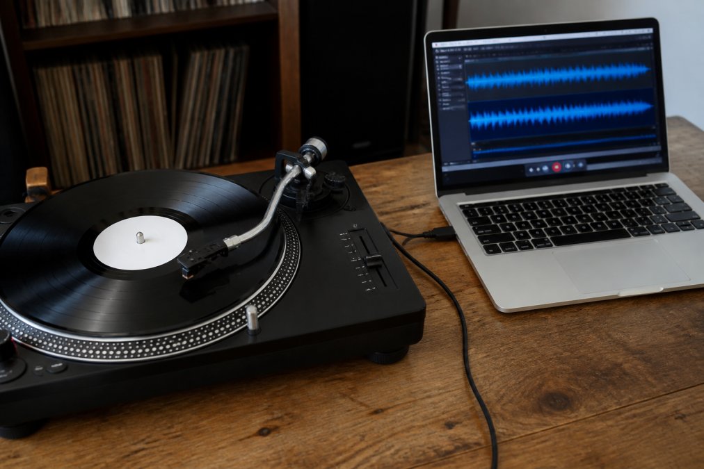 A close-up of a turntable with a vinyl record spinning, connected to a laptop via a USB cable, with recording software open on the screen showing audio waveforms