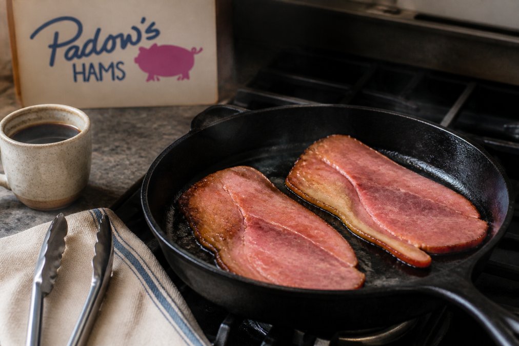 Two country ham slices sizzling in a cast iron skillet with browned edges and a fat cap visible on the stovetop.