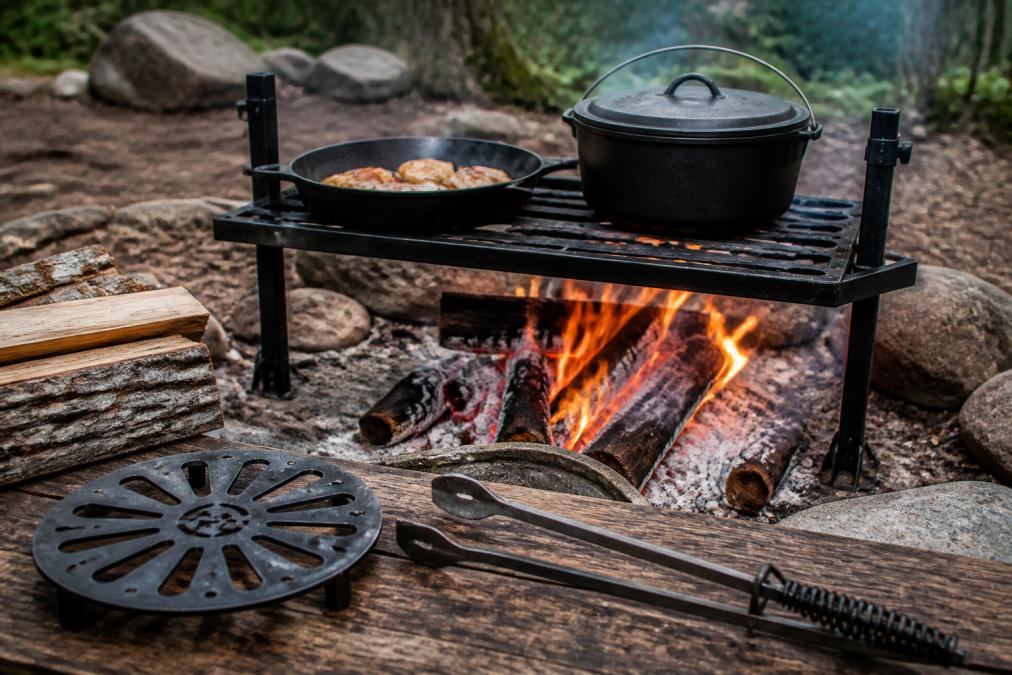 Cast iron skillet and Dutch oven sitting on a heavy-duty adjustable grill grate over an open campfire, surrounded by outdoor cooking tools including tongs and a trivet on a rustic wooden surface