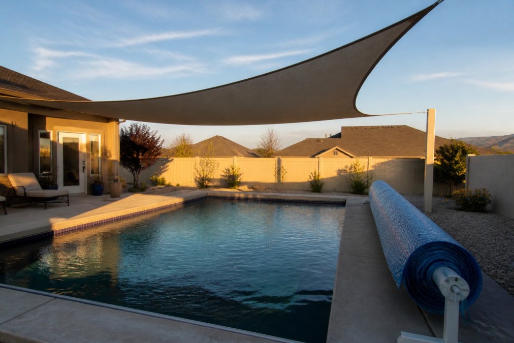 A residential backyard pool with a large triangular shade sail stretched overhead casting a deep shadow across most of the water surface, a pool cover partially rolled back on the concrete deck, late afternoon light