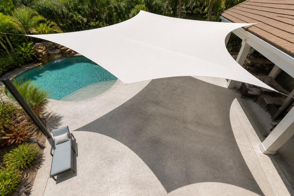 Overhead view of concrete pool deck with white shade sail installed, creating distinct shadow pattern across surface, pool visible in background with tropical landscaping