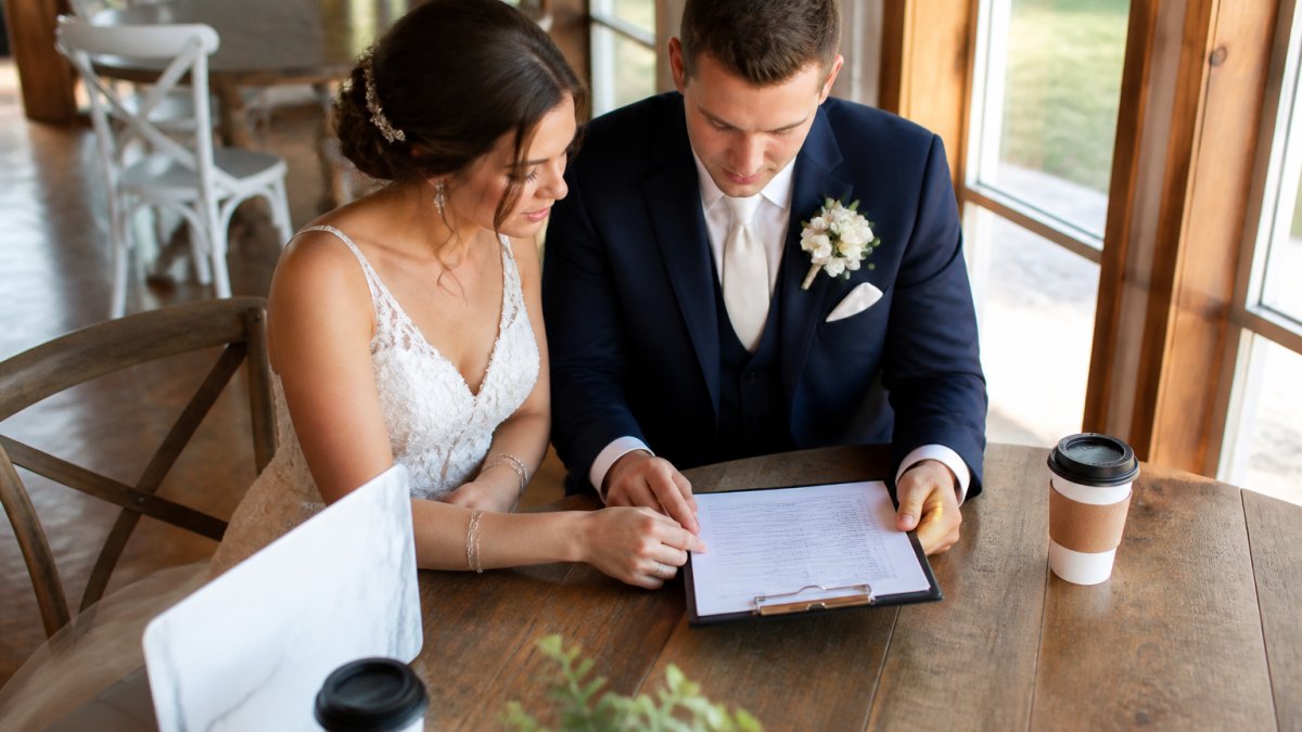A bride and groom reviewing a printed wedding day schedule on a clipboard at a wooden venue table, laptop and two coffee cups nearby, soft afternoon light through tall windows