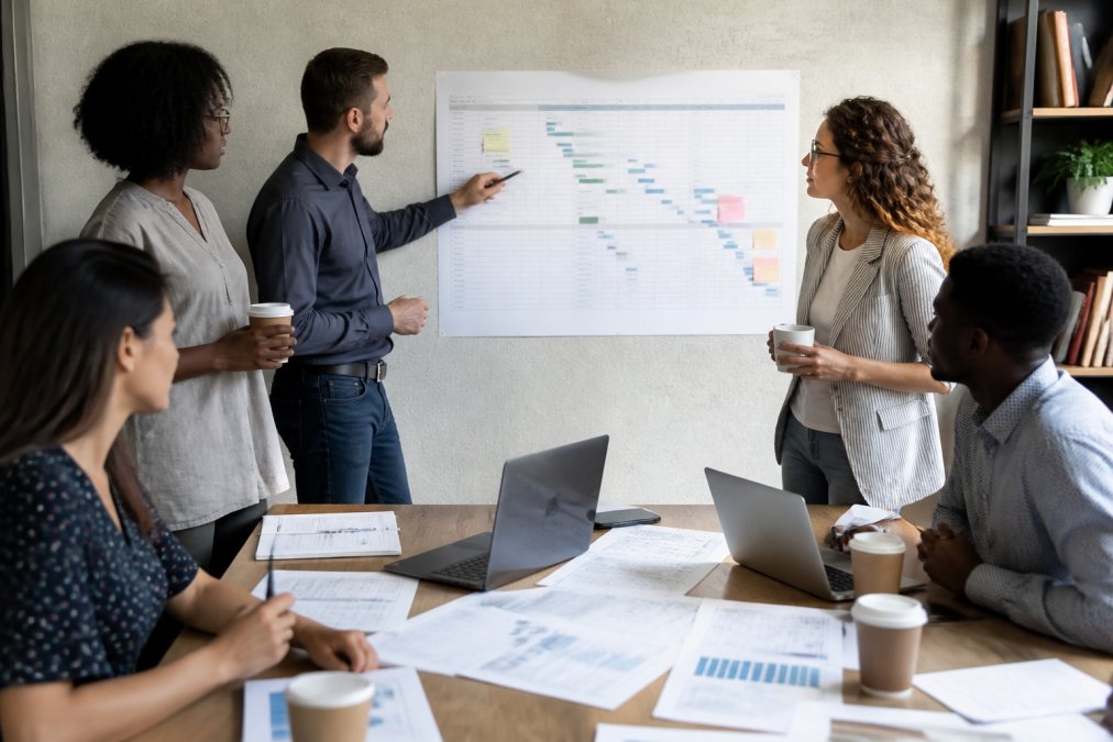 Authors and professionals reviewing a book launch budget timeline and expense spreadsheet on a wall chart