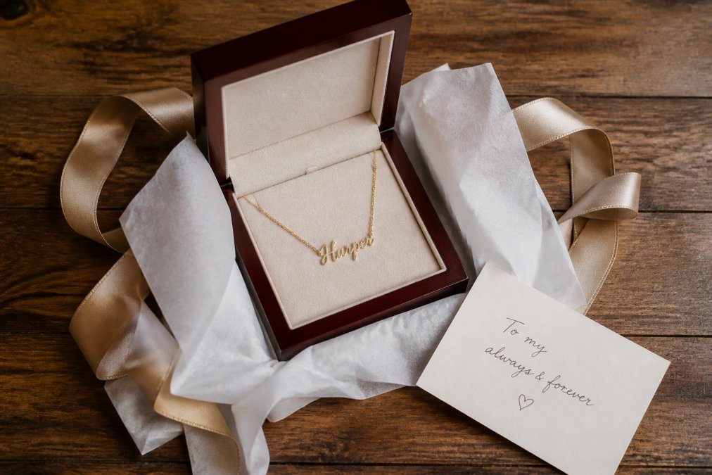 Overhead shot of an open mahogany-style jewelry box with a name necklace inside, soft tissue paper, a handwritten note card, and ribbon on a warm wooden table