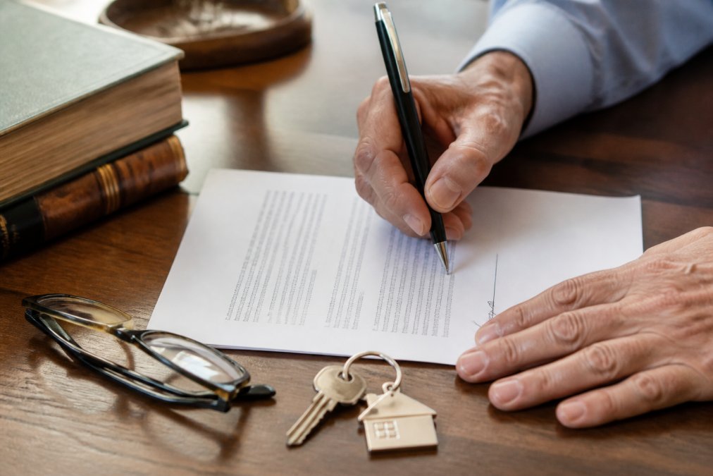 Close-up of hands signing a property deed document on a wooden desk, with a house key and a pair of reading glasses resting nearby in warm indoor lighting
