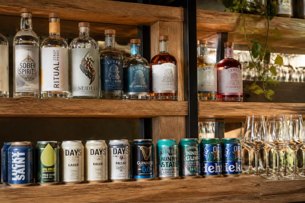 Premium non-alcoholic spirits and alcohol-free beers displayed on a bar shelf with natural lighting, showing bottles and cans with clear labels and professional presentation