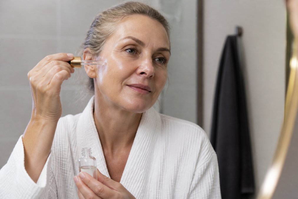 Woman in her 50s applying a hydrating serum to achieve radiant makeup in a serene, softly lit bathroom setting