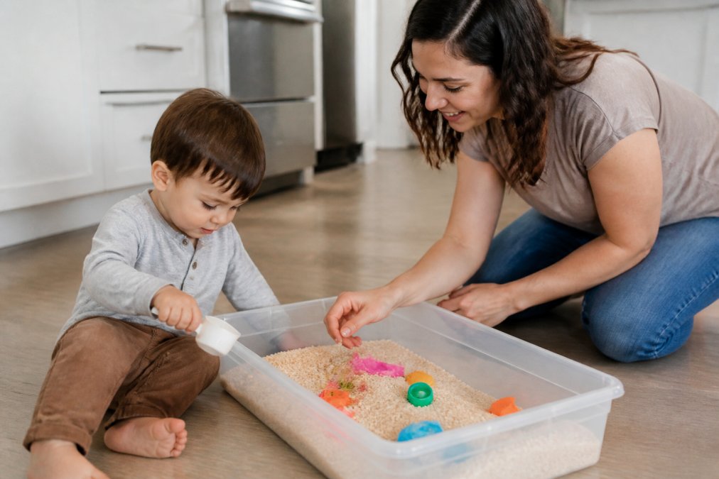 A young child sitting on a kitchen floor exploring a shallow plastic bin filled with dry rice, scoops, and small colorful toys, with a parent kneeling nearby guiding the play in a bright home setting