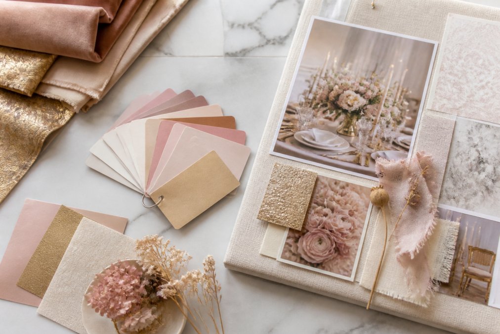 Close-up flat lay of luxury event design materials: fabric swatches in blush and gold tones, printed color palette cards, dried floral samples, and a mood board pinned to a linen board on a marble surface