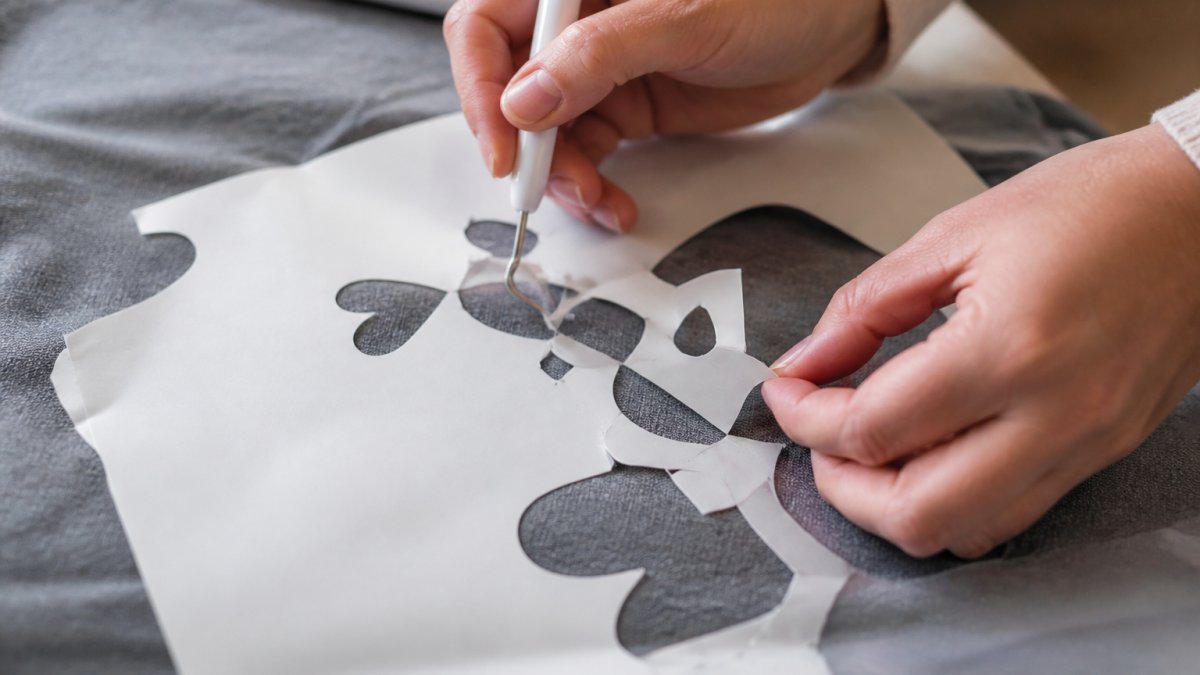 Close-up of hands carefully weeding heat-transfer vinyl from a design template, showing the detail work required for clean application