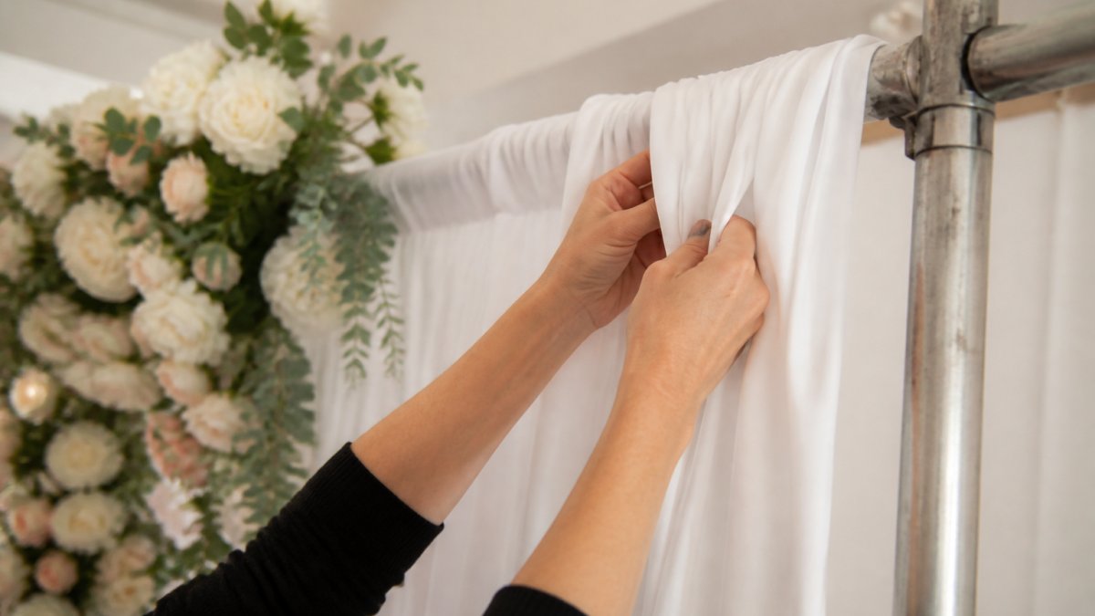 Close-up of hands gathering white fabric into soft pleats on a wedding backdrop frame, with floral accents visible in the background