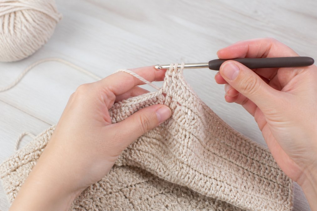Hands working on a partially completed baby sweater, showing crochet hook pulling yarn through loops with completed stitches visible on the hook and finished rows below on a light-colored crafting table