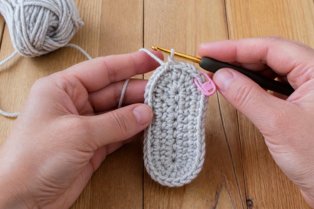 Close-up of a crafter's hands holding a crochet hook and a partially worked baby bootie sole in soft worsted weight yarn, with a stitch marker visible on a bright wooden table