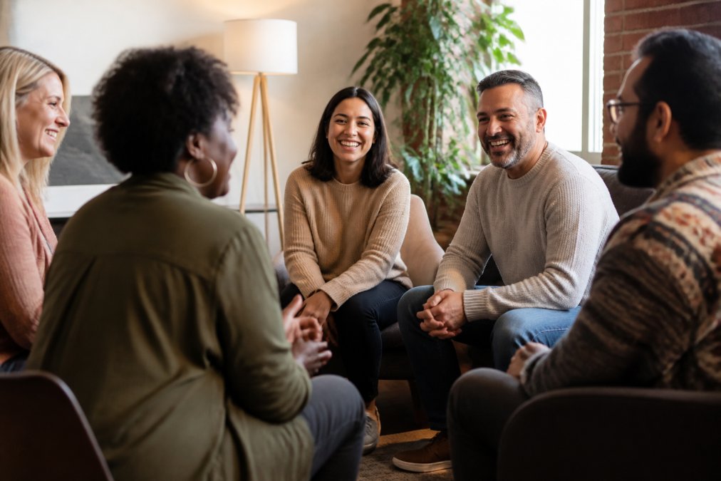 Small group of diverse people sitting in a circle in a warm, welcoming living room space with soft lighting, smiling and engaged in genuine conversation, creating an atmosphere of connection and trust