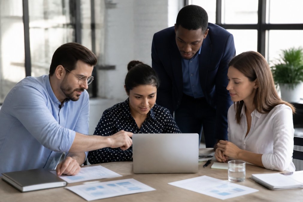 Team members gathered around laptop reviewing business analytics dashboard with growth metrics displayed on screen in collaborative office setting with natural lighting