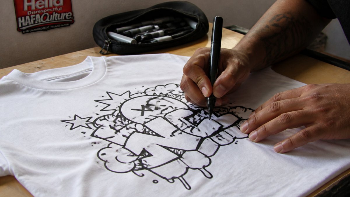 Close-up of hands drawing a bold doodle design on a plain white T-shirt with black fabric markers, showing detailed linework and emerging graphic