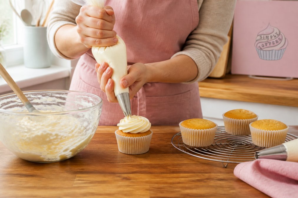 Close-up of a home baker's hands holding a piping bag, piping a swirl of buttercream onto a cupcake on a wooden kitchen counter, soft daylight from a window