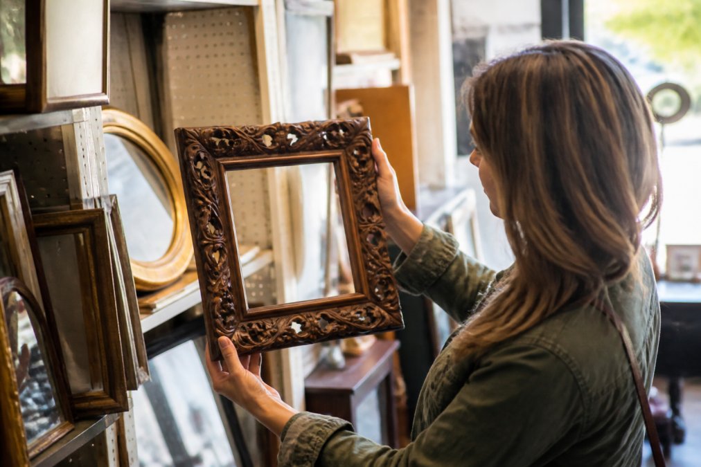 Person browsing and examining vintage decorative frames and mirrors at a thrift store, holding up a ornate wooden frame to inspect it under warm natural lighting