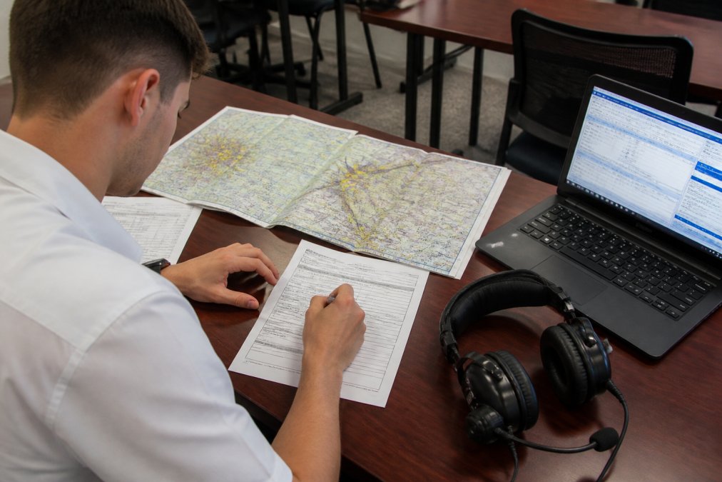 An aviation student seated at a professional training desk filling out FAA flight plan forms, with a sectional chart spread open, a headset resting on the desk, and a laptop displaying flight service software, under bright overhead lighting in a classroom environment