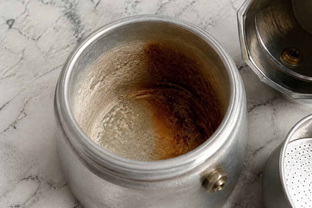 Interior of a moka pot lower chamber showing visible white mineral deposits on one side and brown coffee oil residue on the other, with natural lighting