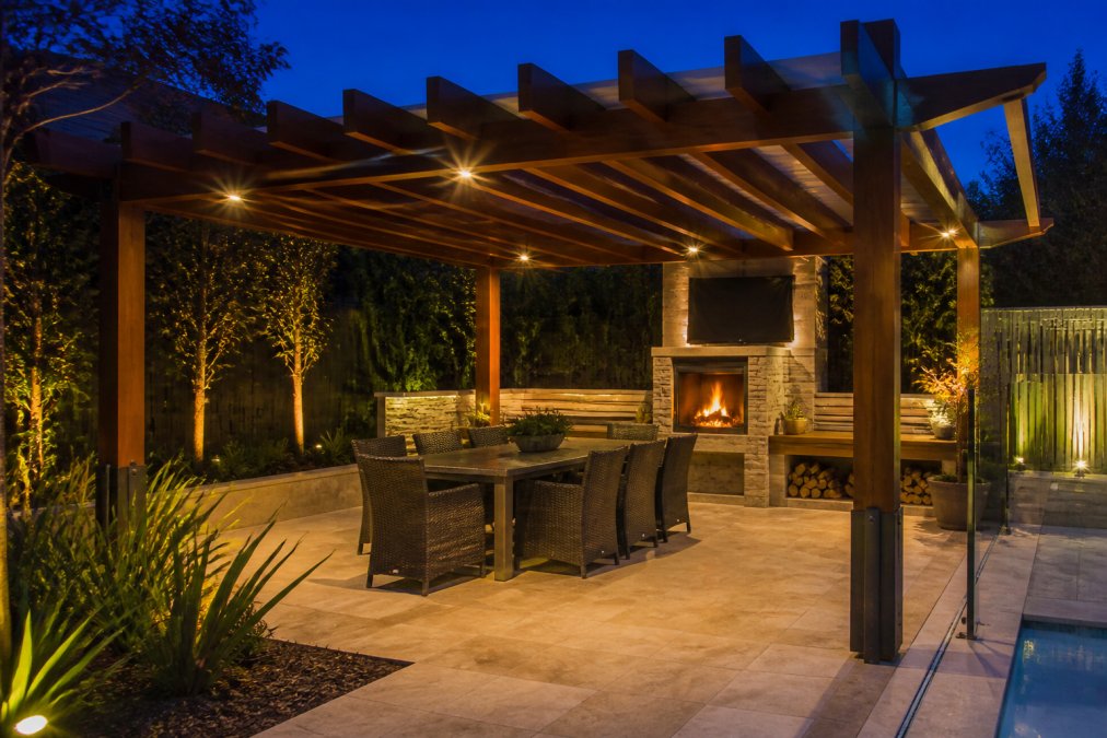 Evening view of a completed alfresco entertaining area with warm ambient lighting, timber pergola providing shade structure, and outdoor dining furniture arranged for entertaining guests