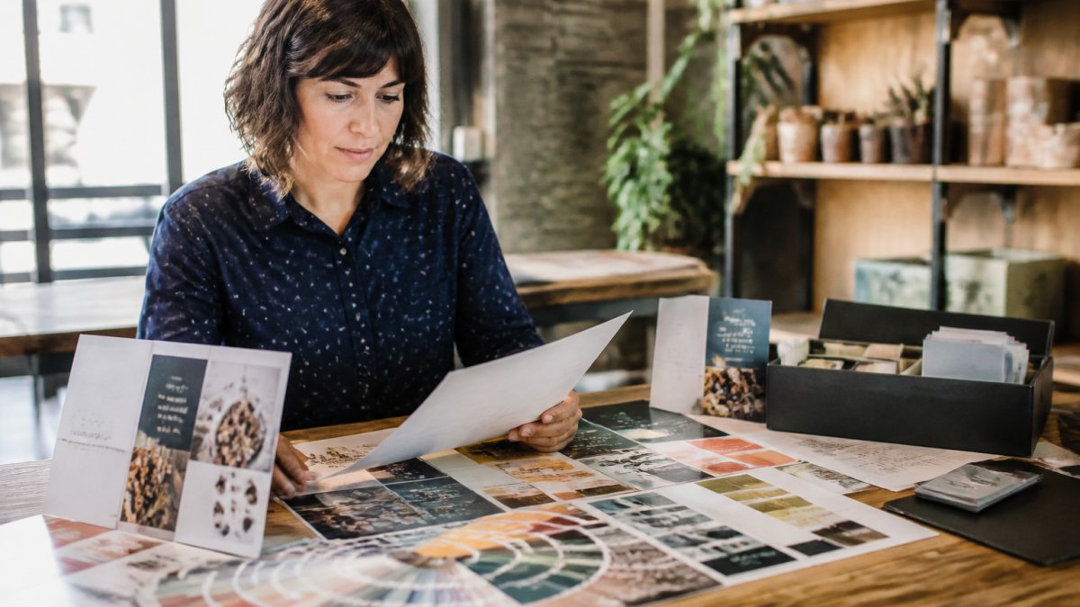Marketing professional reviewing brand guidelines document with printed POS material samples spread across desk, color swatches and typography samples visible under warm office lighting