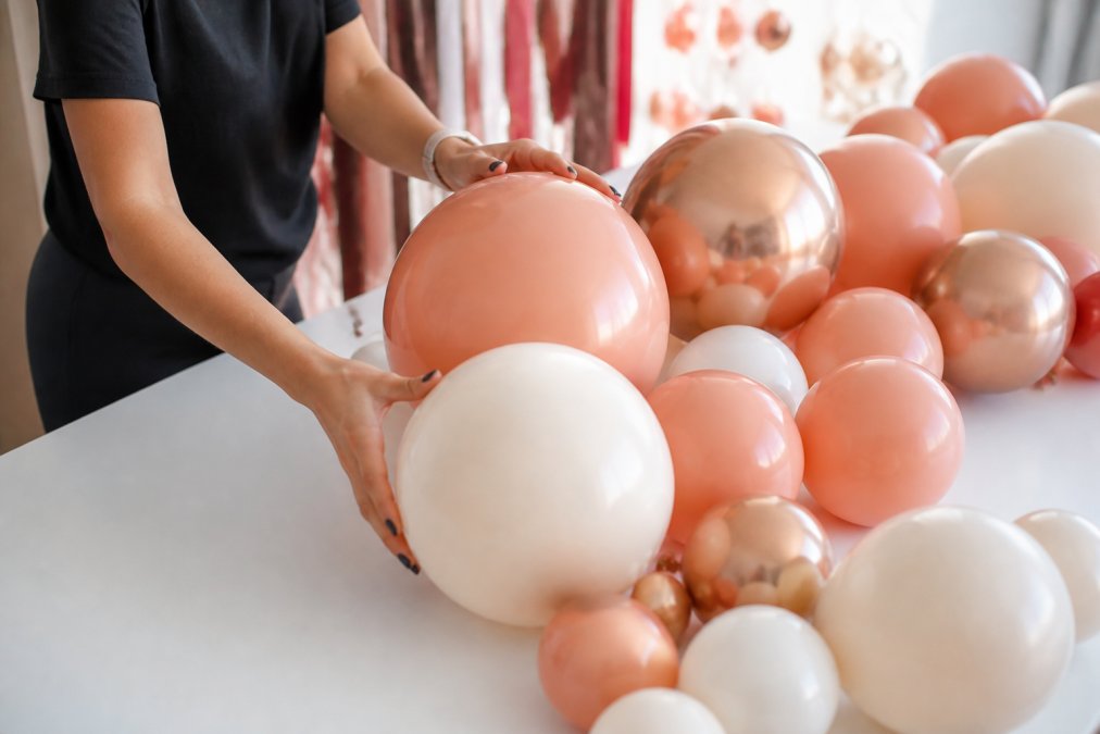 Close-up of hands arranging latex balloons in coordinating colors on a white table, testing different sizes and color combinations together under bright natural lighting