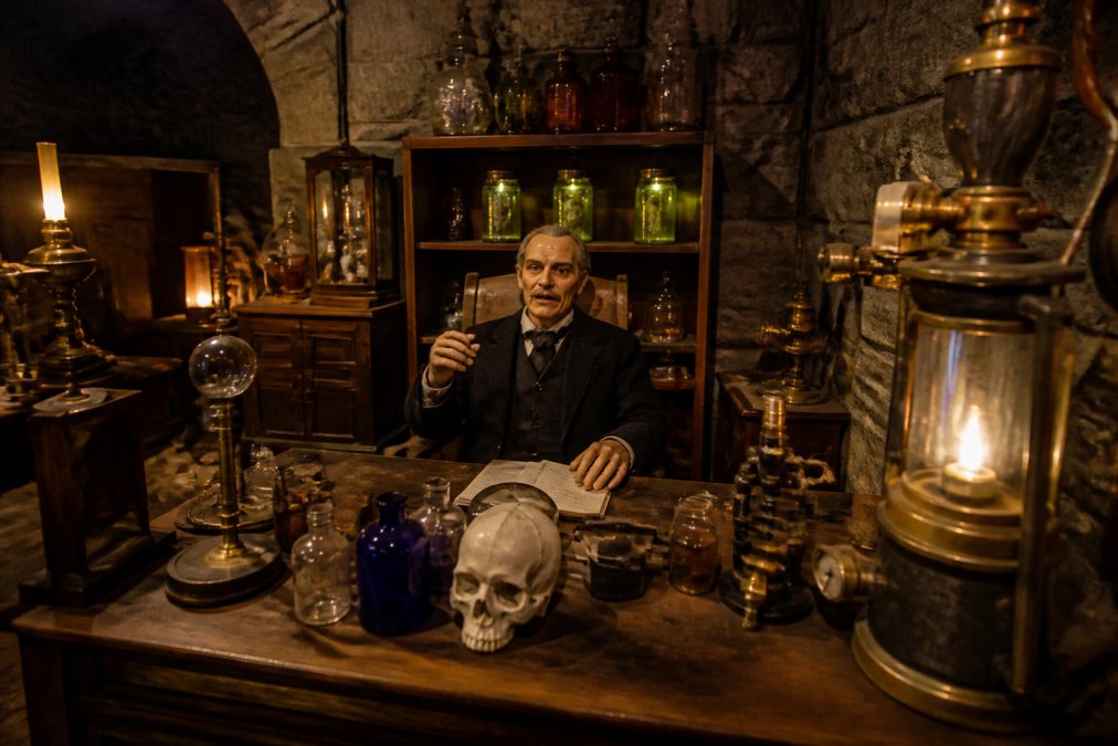 A dimly lit escape room interior with vintage laboratory props, glowing jars, brass instruments, and an animatronic figure positioned behind a desk, creating a gothic steampunk atmosphere