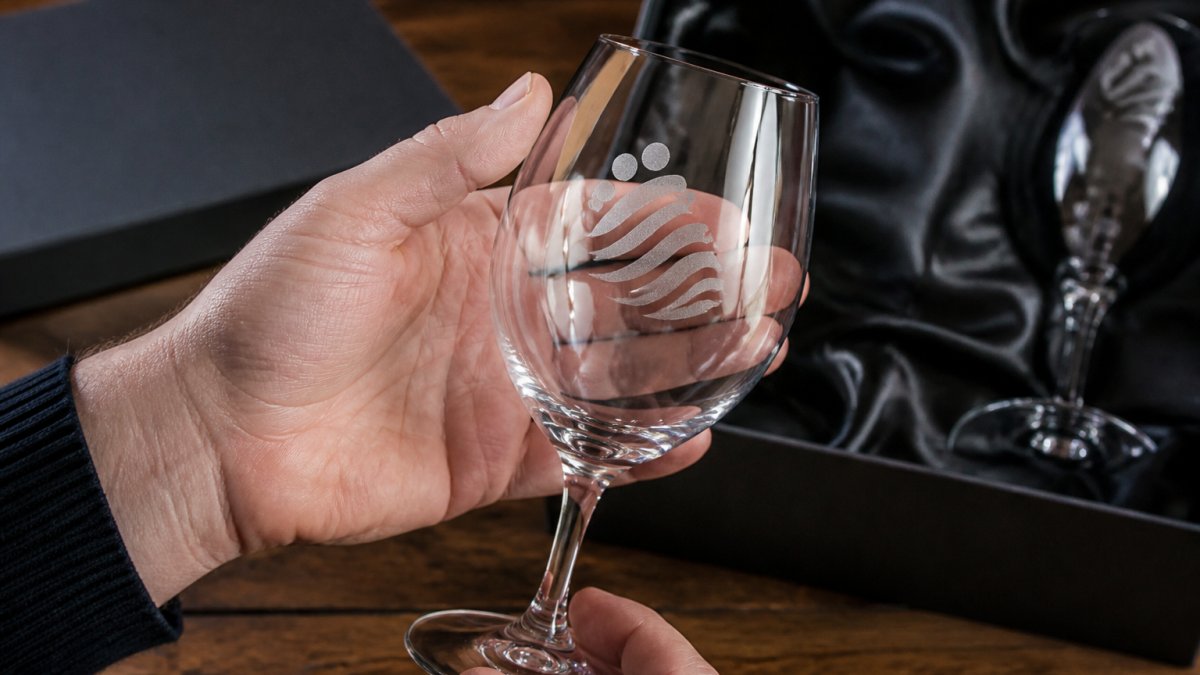 Close-up of hands holding a clear etched wine glass against soft studio light, showing a frosted logo positioned on the curved bowl with crisp edges and visible contrast