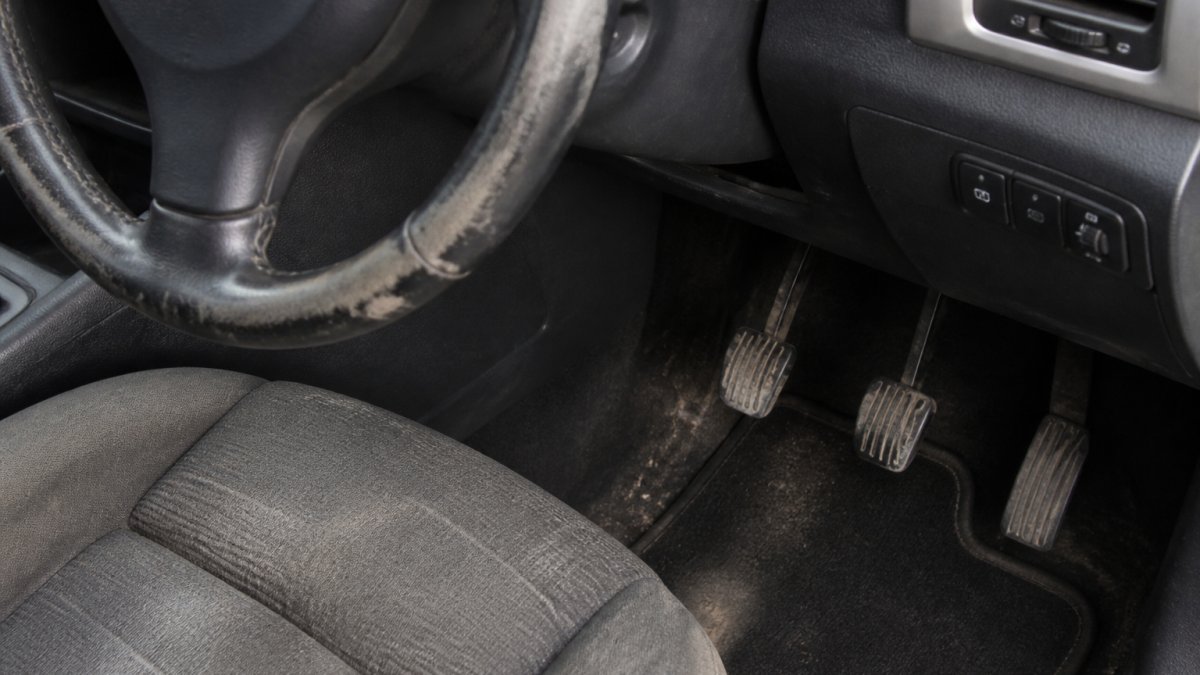 Close-up of worn car interior showing steering wheel grip deterioration, heavily scuffed pedals with rubber worn away, and aged seat fabric with visible creasing and fading in natural daylight