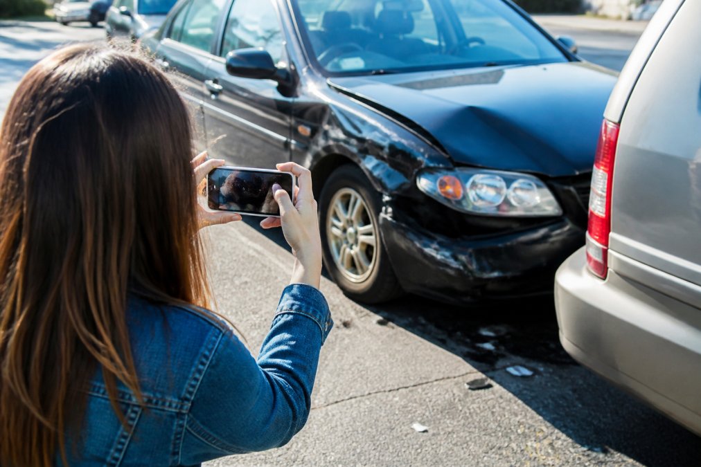 Passenger in a rideshare vehicle documenting accident scene with smartphone camera, capturing vehicle damage and surrounding street context with natural daylight