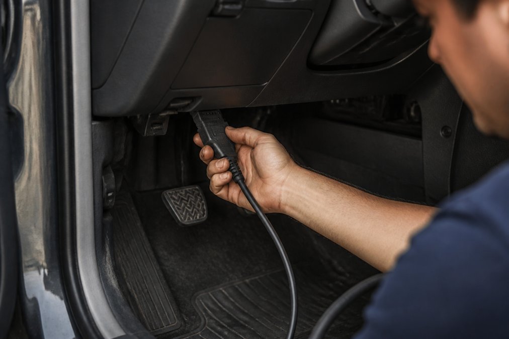 Professional technician's hands connecting an OBD-II scanner cable into the data link connector under a vehicle dashboard with natural workshop lighting