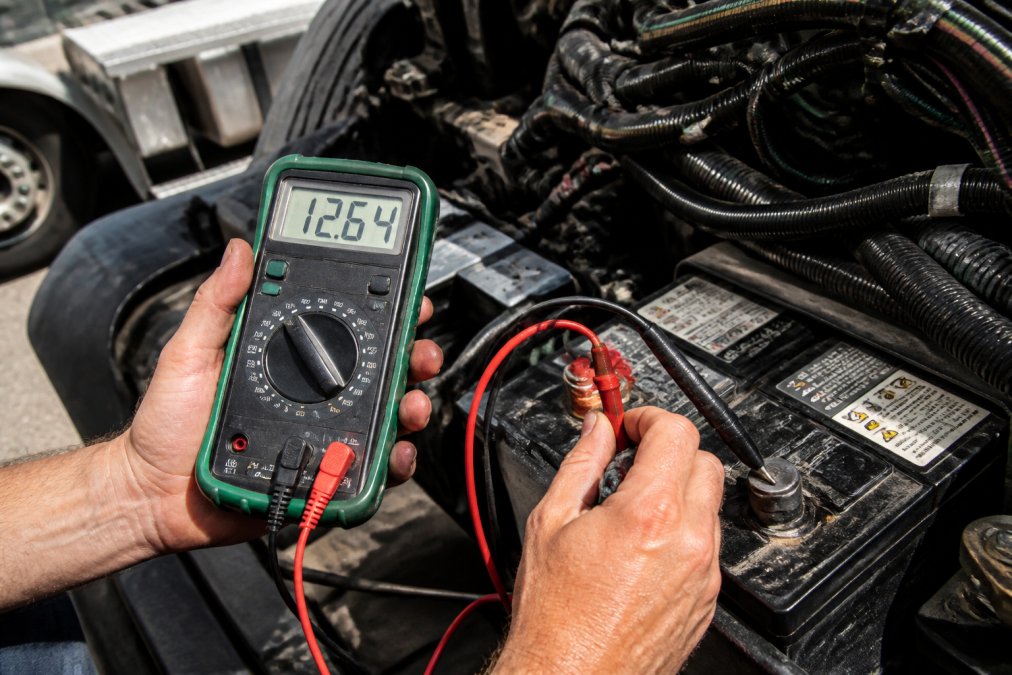 Technician's hands holding a digital multimeter with red and black probes positioned against a semi truck battery terminal, showing proper probe placement and the meter display reading voltage in a bright outdoor setting