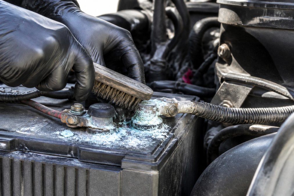 Close-up of a technician's gloved hands using a wire brush to scrub heavy white and blue corrosion buildup from a semi truck battery terminal, with visible terminal oxidation and corroded cable connection in natural daylight