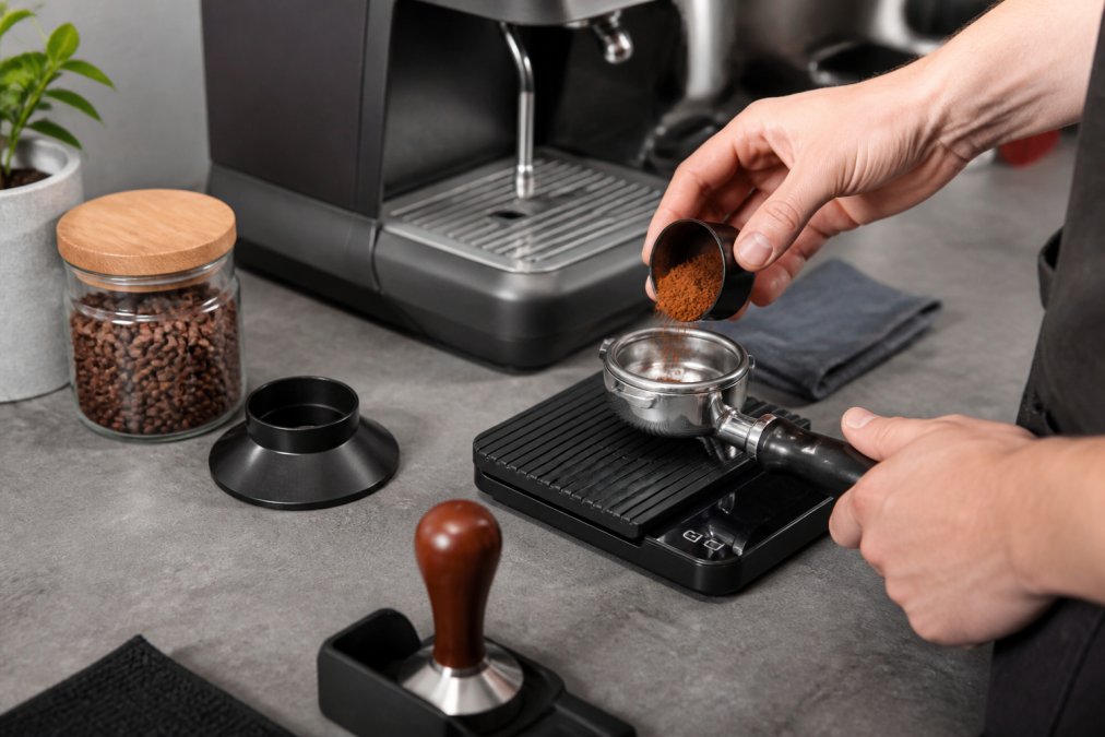 A home barista's hands weighing ground coffee in a portafilter on a digital scale, with a timer and espresso machine in the background on a kitchen counter