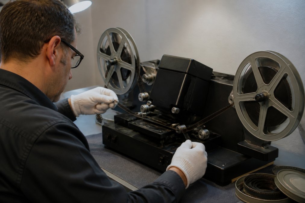 Professional technician carefully threading aged 8mm film through a professional digitization scanner in a climate-controlled studio, with film reels positioned on either side and soft studio lighting illuminating the equipment
