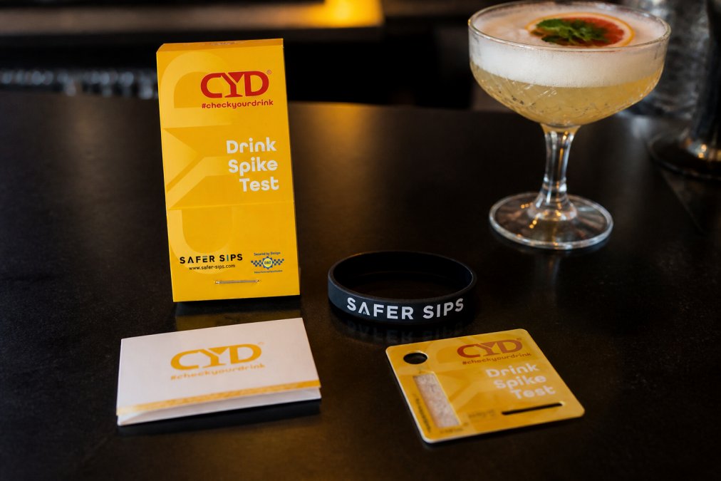 Flat lay of several drink spiking test strip products, a wristband, a matchbook-style strip pack, and a card-style test, arranged on a dark surface alongside a cocktail glass with ambient bar lighting