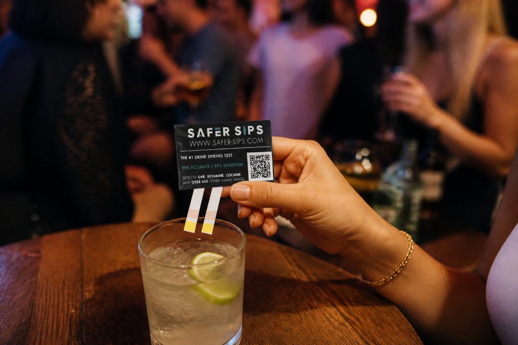 Close-up of a person's hand discreetly holding a small drink testing strip over a glass at a dimly lit bar, other people socialising out of focus in the background, warm ambient lighting