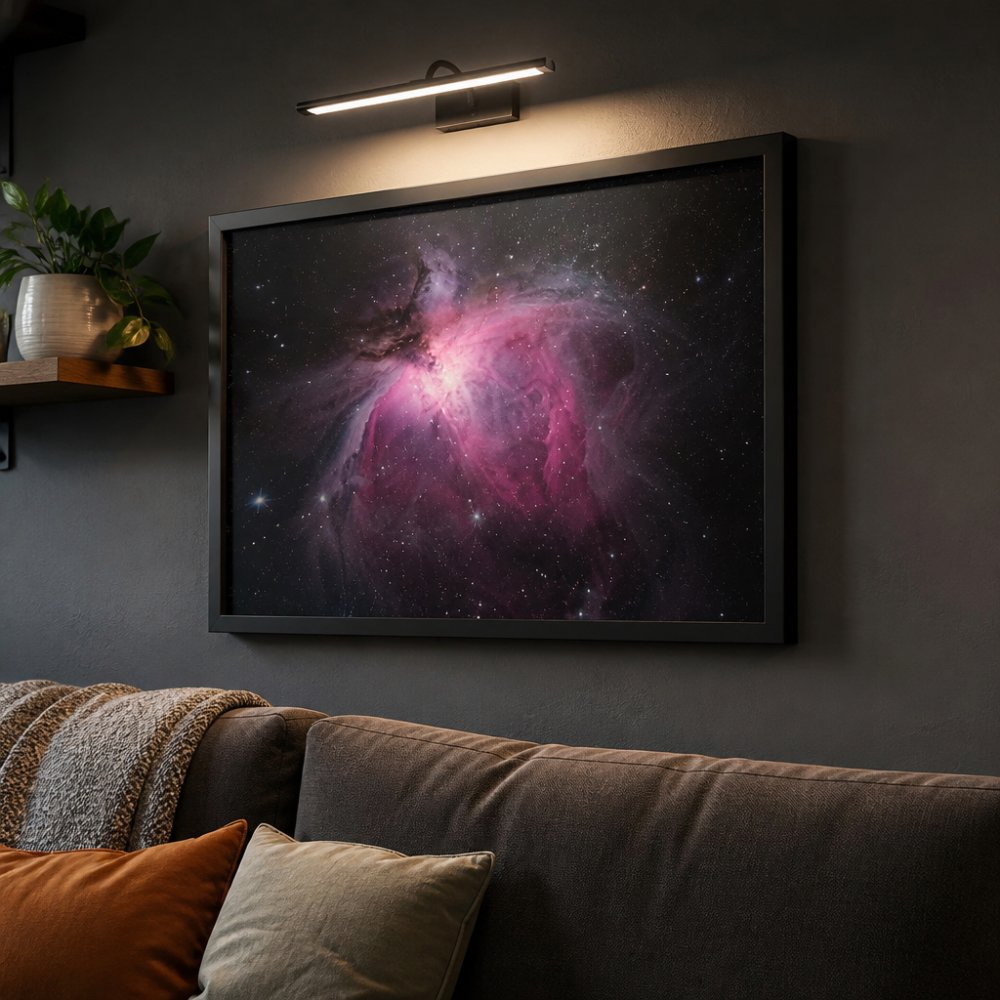 A framed astrophotography print of the Orion Nebula mounted on a dark gray living room wall, illuminated by a small adjustable picture light angled from above, with no visible glare on the glass