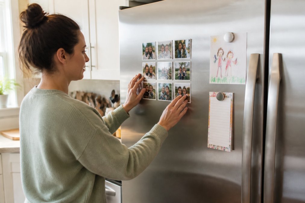 A parent arranging family photos on fridge doors in a neat grid pattern within a bright kitchen.