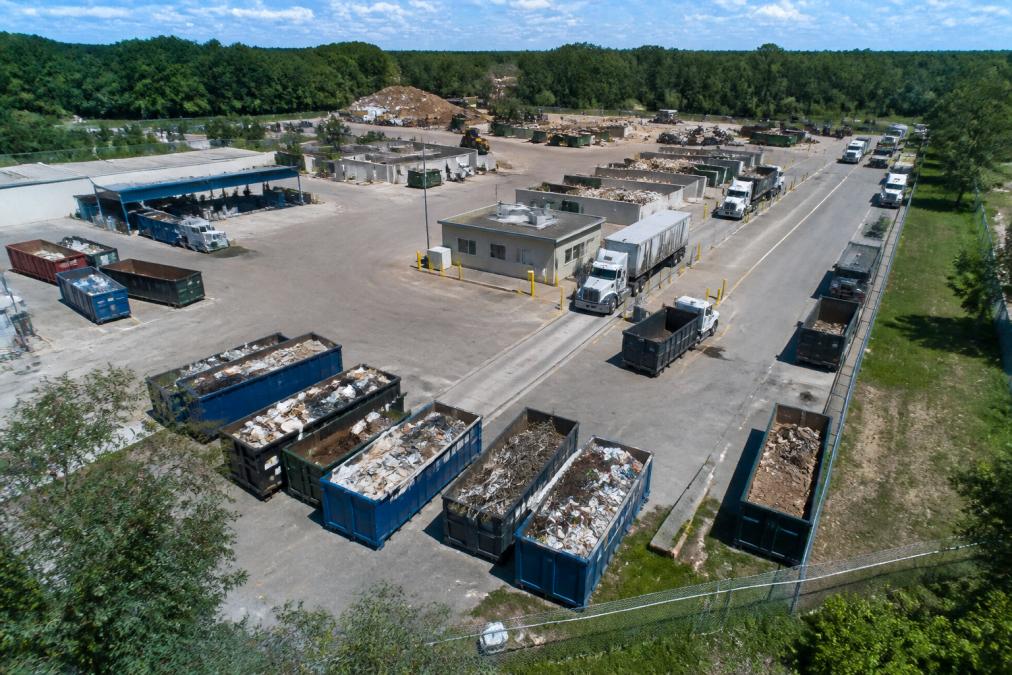 An aerial view of a busy transfer station with multiple roll-off containers, a scalehouse building, and trucks lined up for inspection in bright daylight